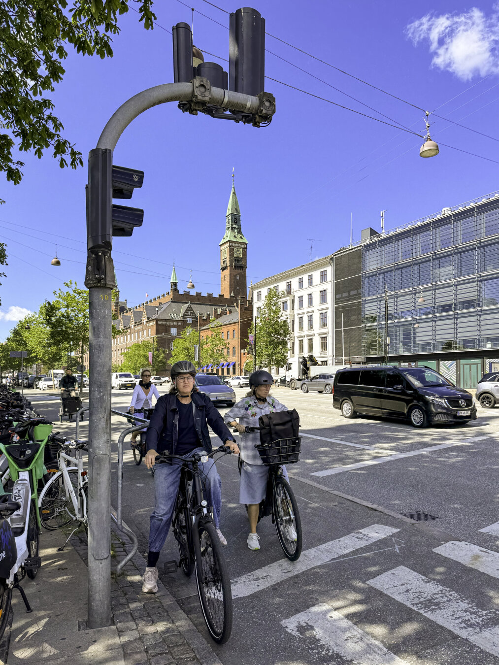 Bicyclists wait at an intersection next to a traffic light that hangs above them and visually spans over a tall historic clock tower in the distance.