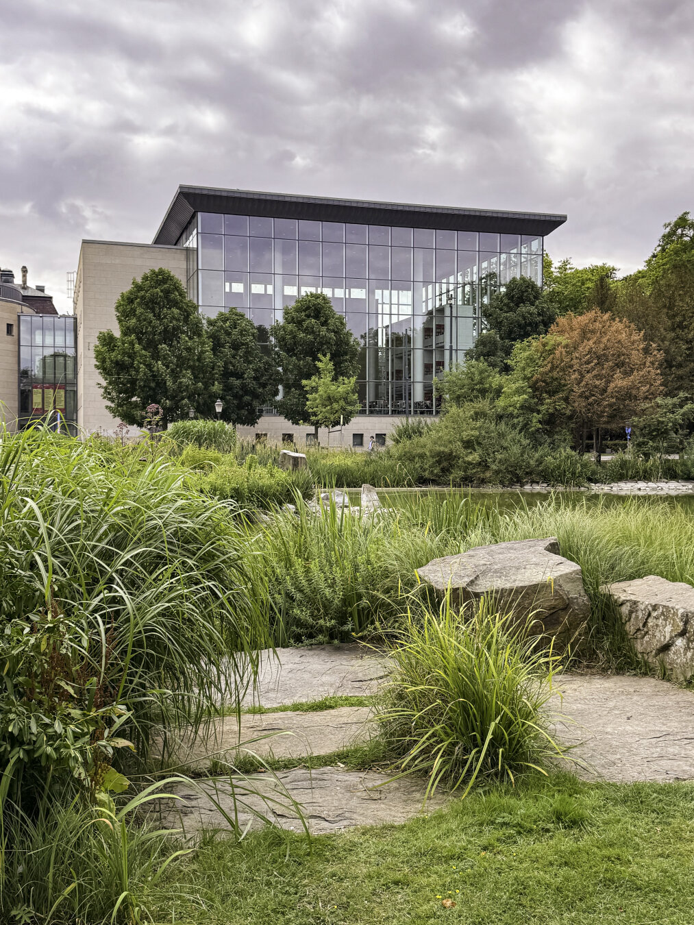 A modernist building with glass façades overlooks a pond with lush vegetation surrounding it.