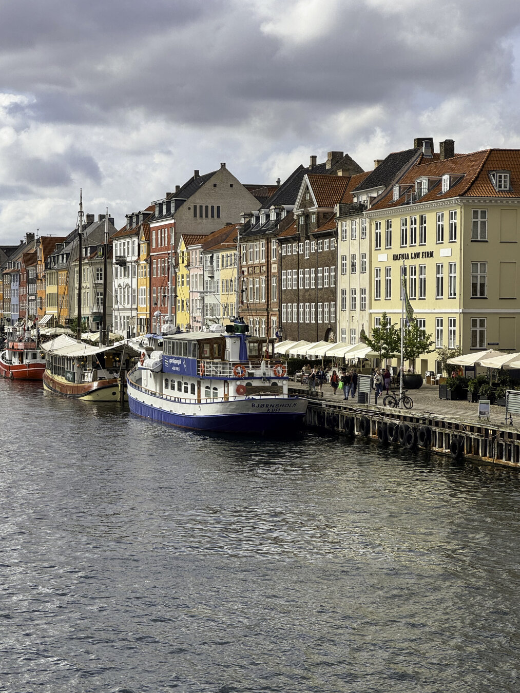 Colorful building façades create a streetwall along a canal with a vibrant pedestrian walkway full of vendor tents.