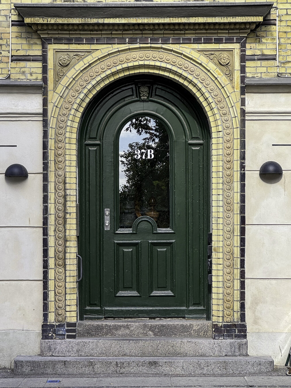 Green door surrounded by a decorative arch comprised of concentric rings of yellow terra cotta tiles, framed by a brown-brick outline.