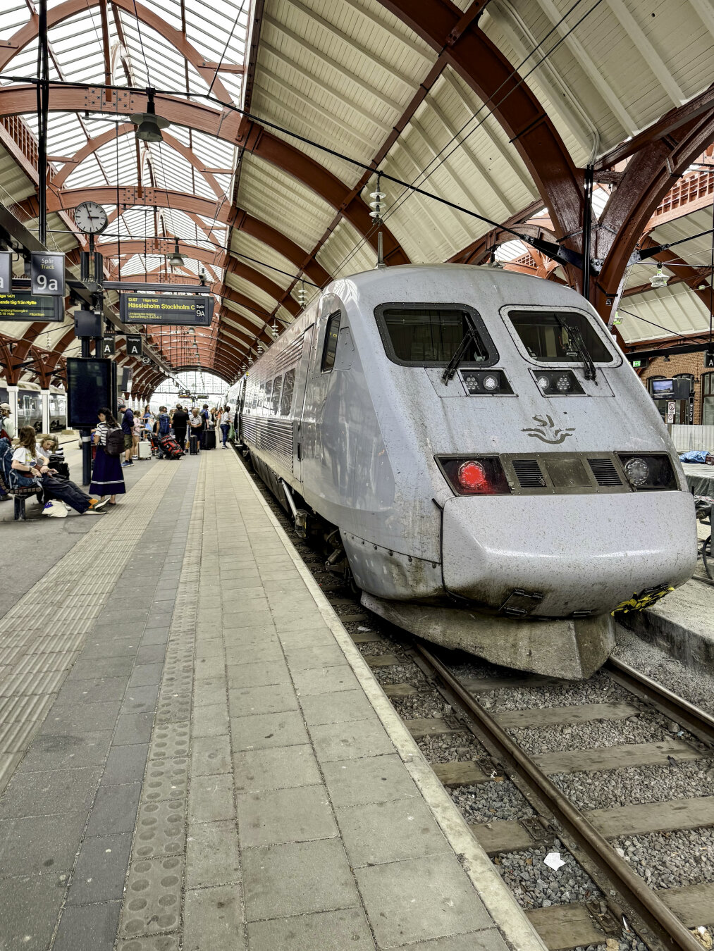 Front of a gray high-speed train sitting at the boarding platform under a red-and-white vaulted ceiling with passengers queued waiting to board.