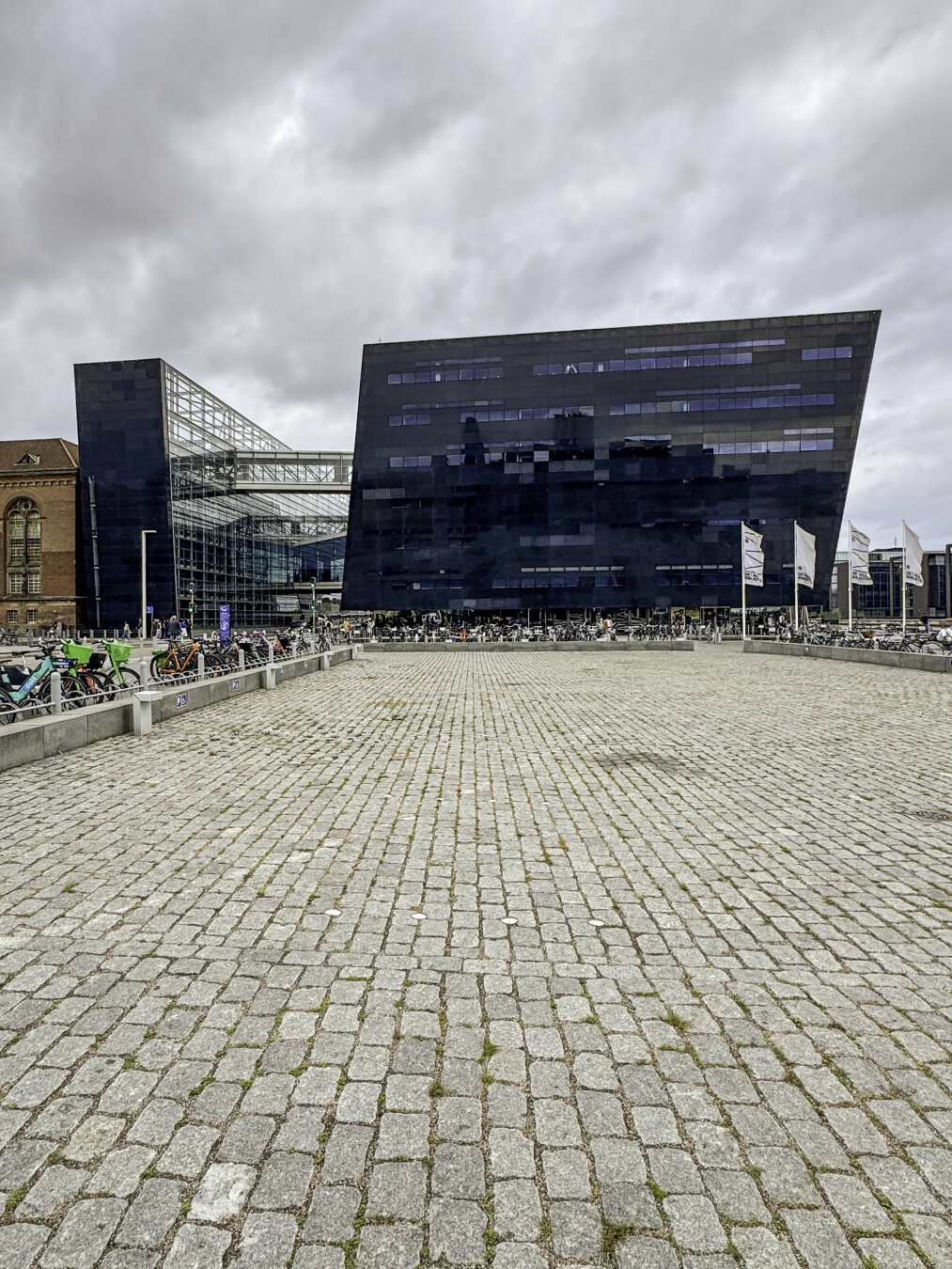 A black-glass modernist building connects via skybridges across a street to an atrium of similar design, attached to a brick historic building. In the foreground, a huge plaza covered in brick pavers holds hundreds of bicycle racks.