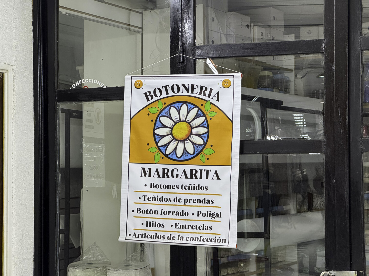 Banner sign hanging on a storefront window that reads "Botonería" with a large daisy inside an orange frame.