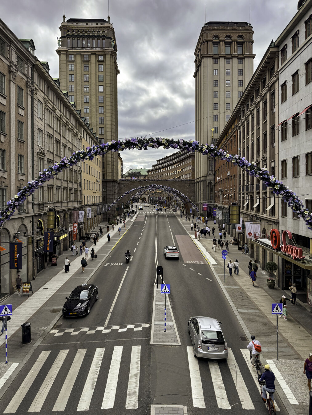 View from a bridge over a busy city street looking toward another bridge a block away framed by two historic towers. Flowered arches span across the street.