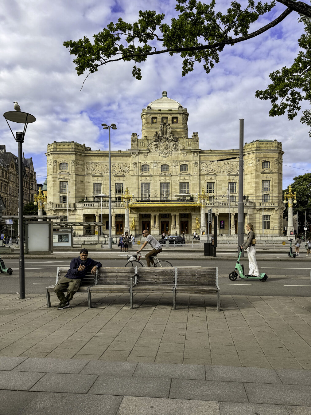 View of a historic building with, in the foreground, a man on a bicycle and a woman on a scooter go by while a man sits on a bench looking at his phone.