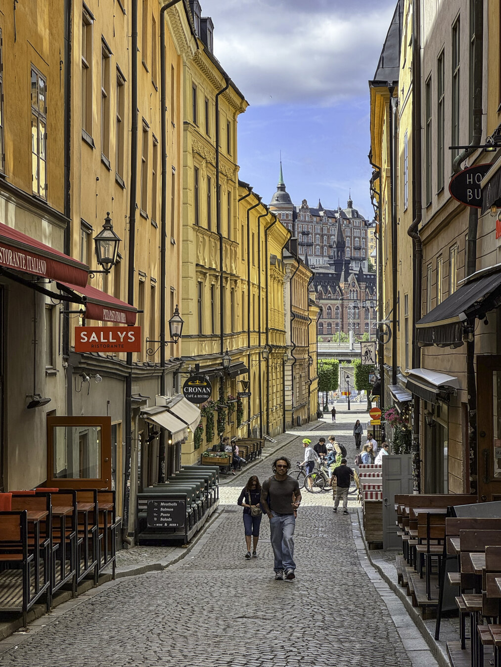 A narrow commercial street is lined with colorful historic building façades, storefronts, and restaurant patios. A few pedestrians and bicyclists walk along it. Larger historic urban buildings are in the distance.