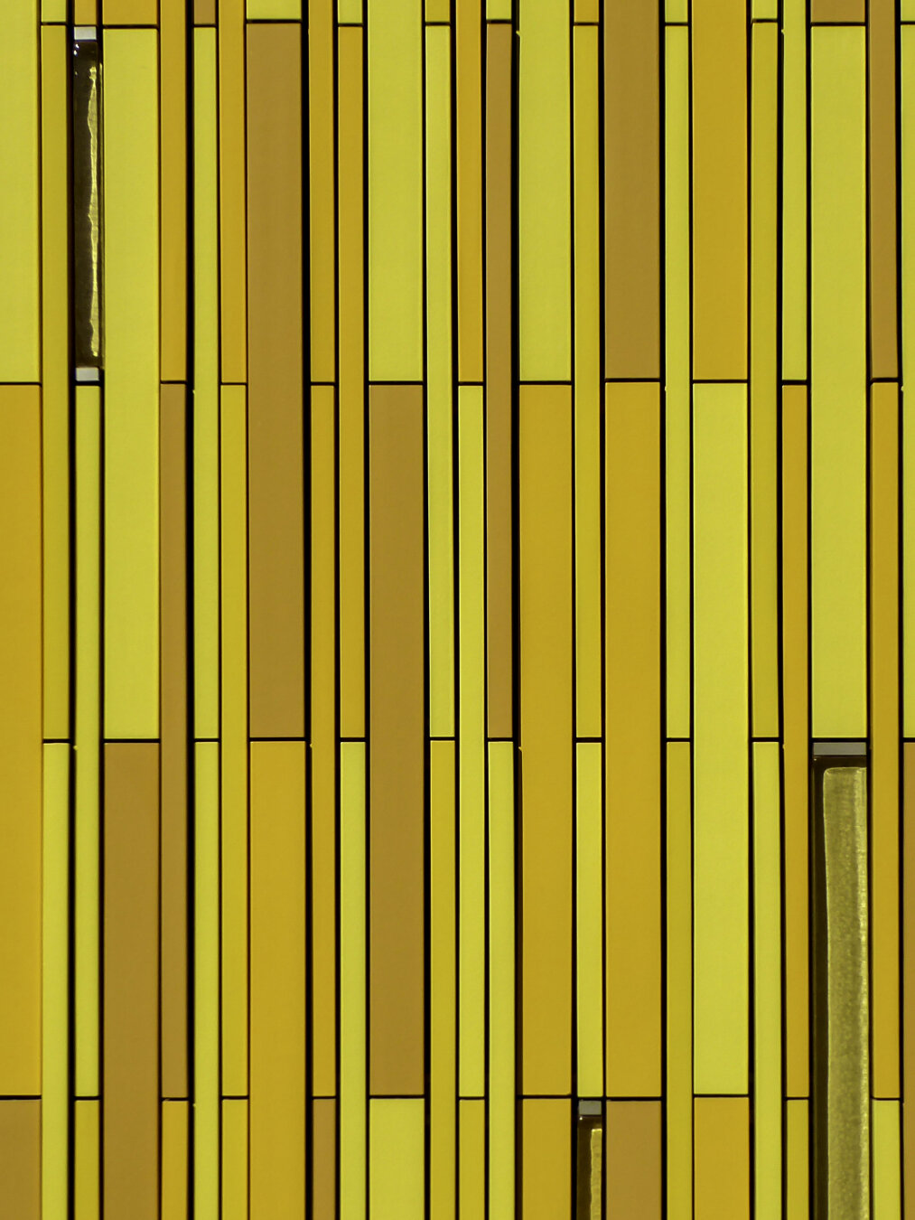 Close-up view of a building facade featuring vertical terra cotta bars in a variety of golds and yellows with narrow shadowy gaps in between.