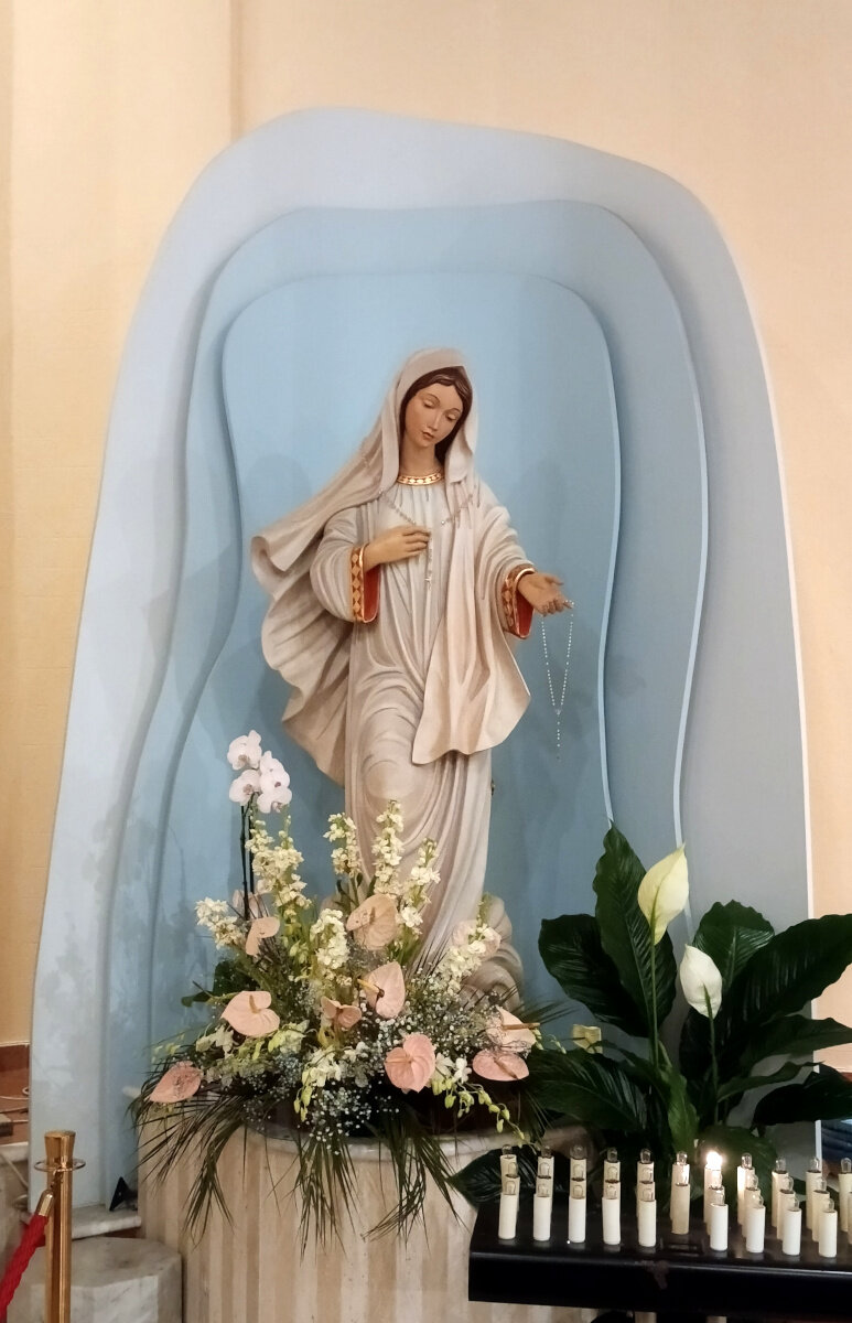 Statua della Madonna di Medjugorje che si trova nel Tempio di San Luca Evangelista. La statua rappresenta la Madonna in piedi, vestita di bianco, con la mano destra al petto e la sinistra in avanti col palmo all'insù. Il fondale è formato da tre stradi sfalsati di sfumature di azzurro. Ai suoi piedi ci sono dei cesti con fiori.