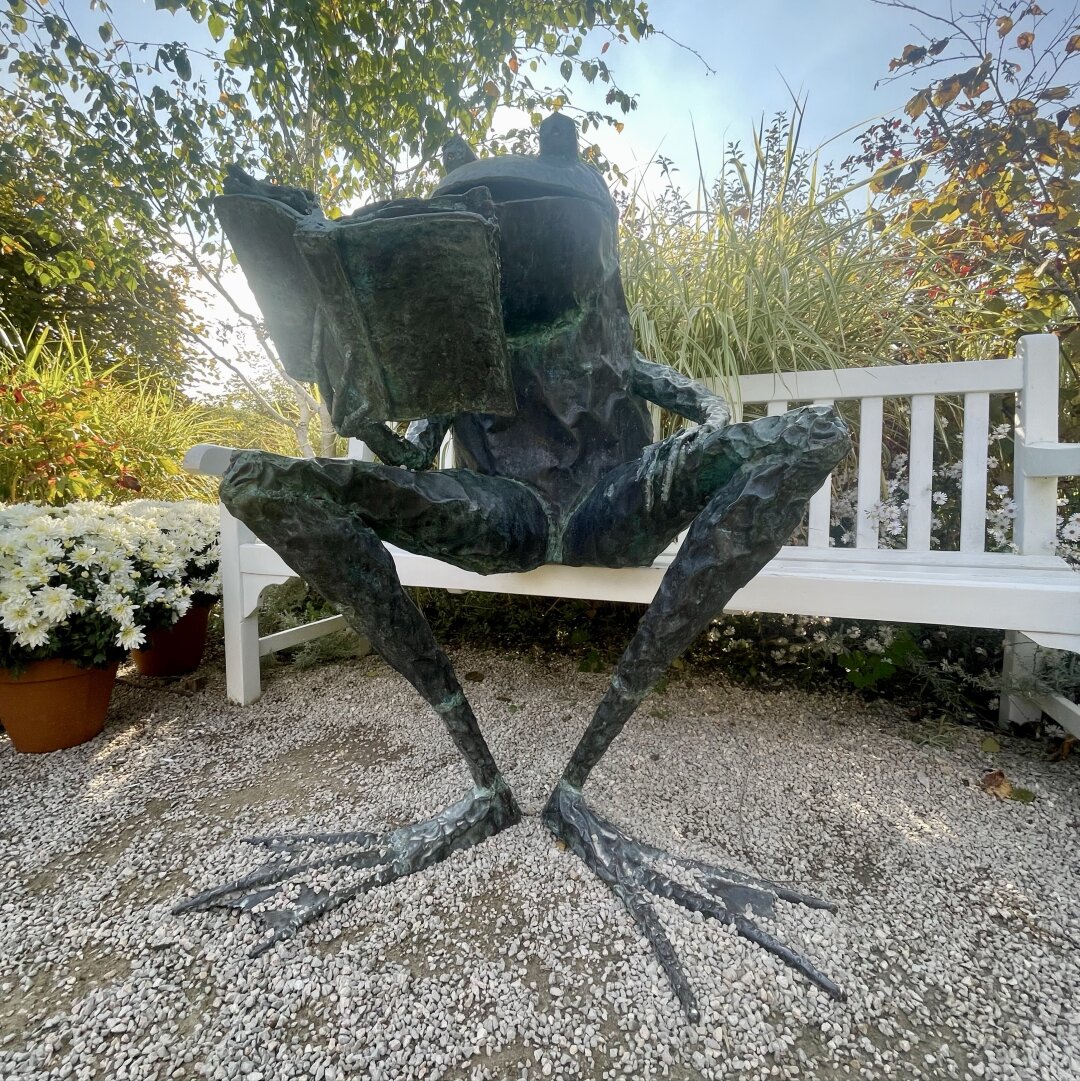 Sculpture en acier vert de grenouille lisant assise sur un banc. Taille humaine.