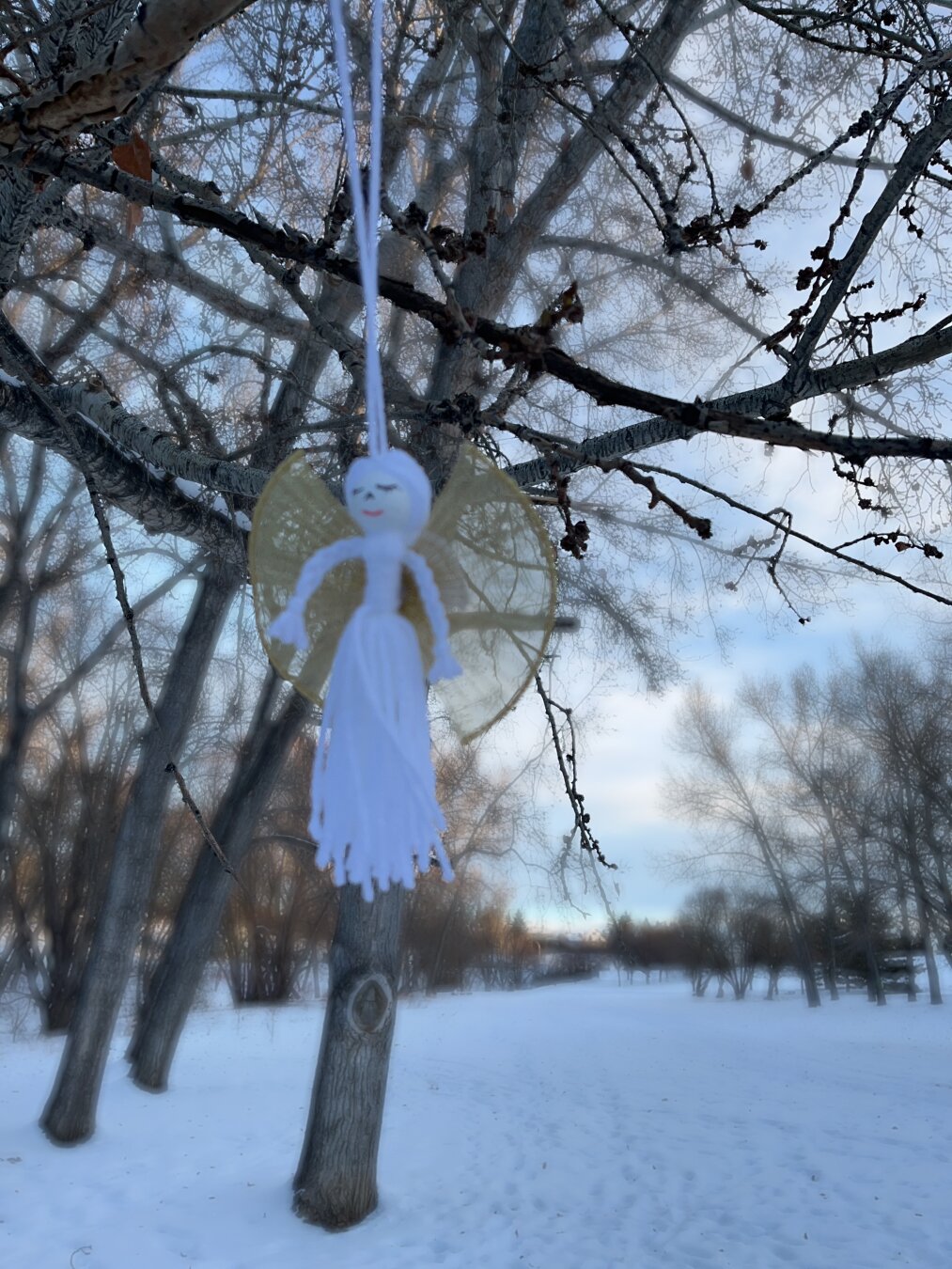 A small angel ornament hangs from a tree branch in a snowy landscape, with a cloudy sky and leafless trees in the background.