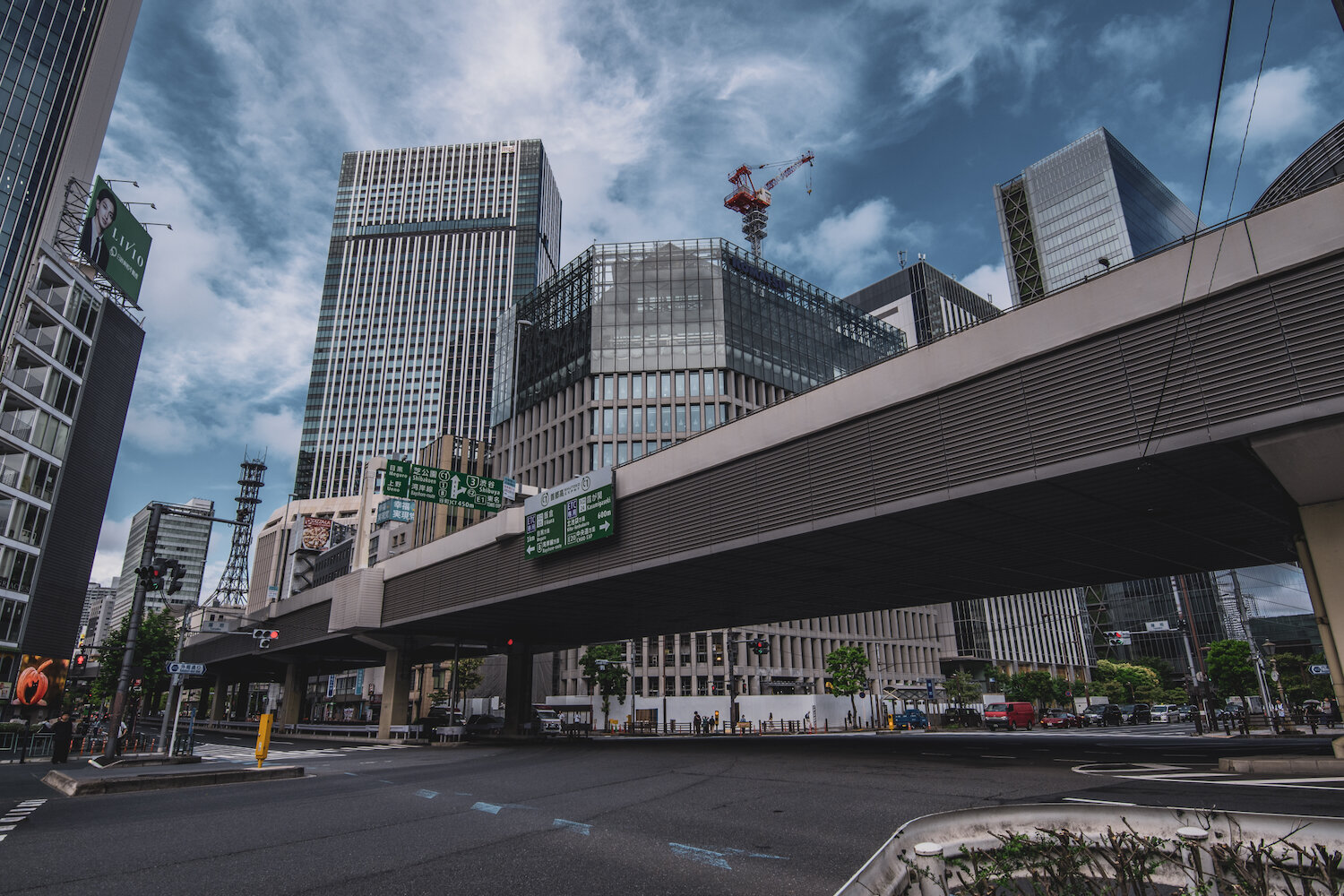 An elevated highway stretches horizontally across Tameike junction in Akasaka 2-chome, Minato, Tokyo. Towering modern glass and steel skyscrapers reach towards a dramatic, cloud-filled sky, whilst a bright red construction crane stands atop a structure under development, silently capturing the calm, structured rhythm and constant evolution of this bustling metropolis.