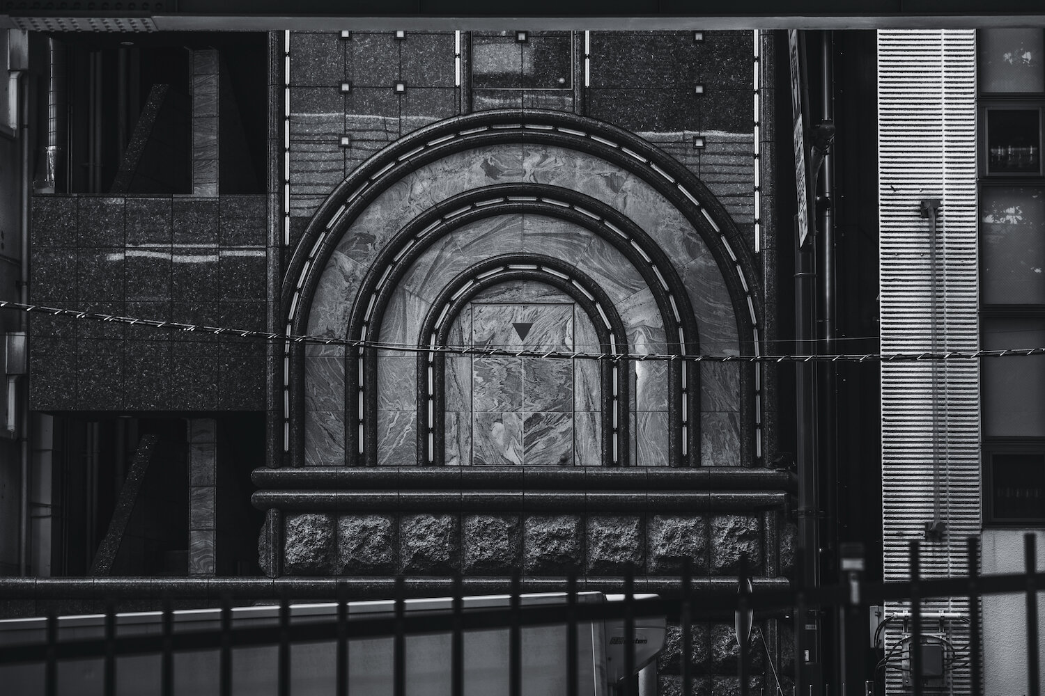 A detailed monochrome photograph captures a portion of a textured urban building facade. Concentric semi-circular stone arch forms dominate the composition, appearing framed by horizontal industrial grid lines. Light accentuates the rich stone textures and patterns, while other complex urban infrastructure lines cross the foreground. It is a moment of architectural contemplation in a busy city.