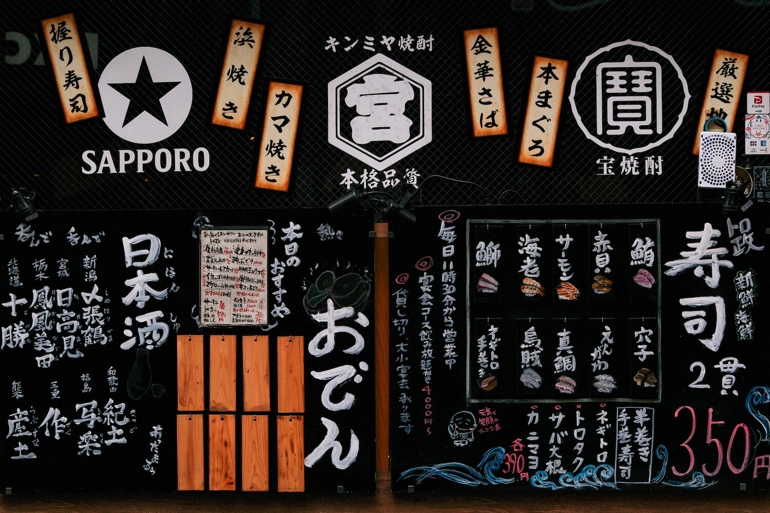 A black izakaya menu board filled with lively Japanese calligraphy listing sake brands, sushi, and oden, beneath glowing brand logos of shochu and beer. The hand-painted signs present a vibrant culinary narrative, evoking the energetic atmosphere of a modern Japanese tavern in full swing.