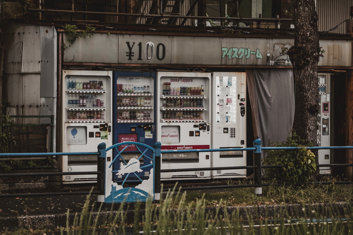 A row of weathered Japanese vending machines stands under a retro shop canopy showing signs of age. Glowing softly with colourful beverages inside, these quiet machines dutifully serve passers-by. Set against rustic textures and muted urban tones, the scene evokes a nostalgic yet reassuring atmosphere of everyday reliability in a quiet corner.