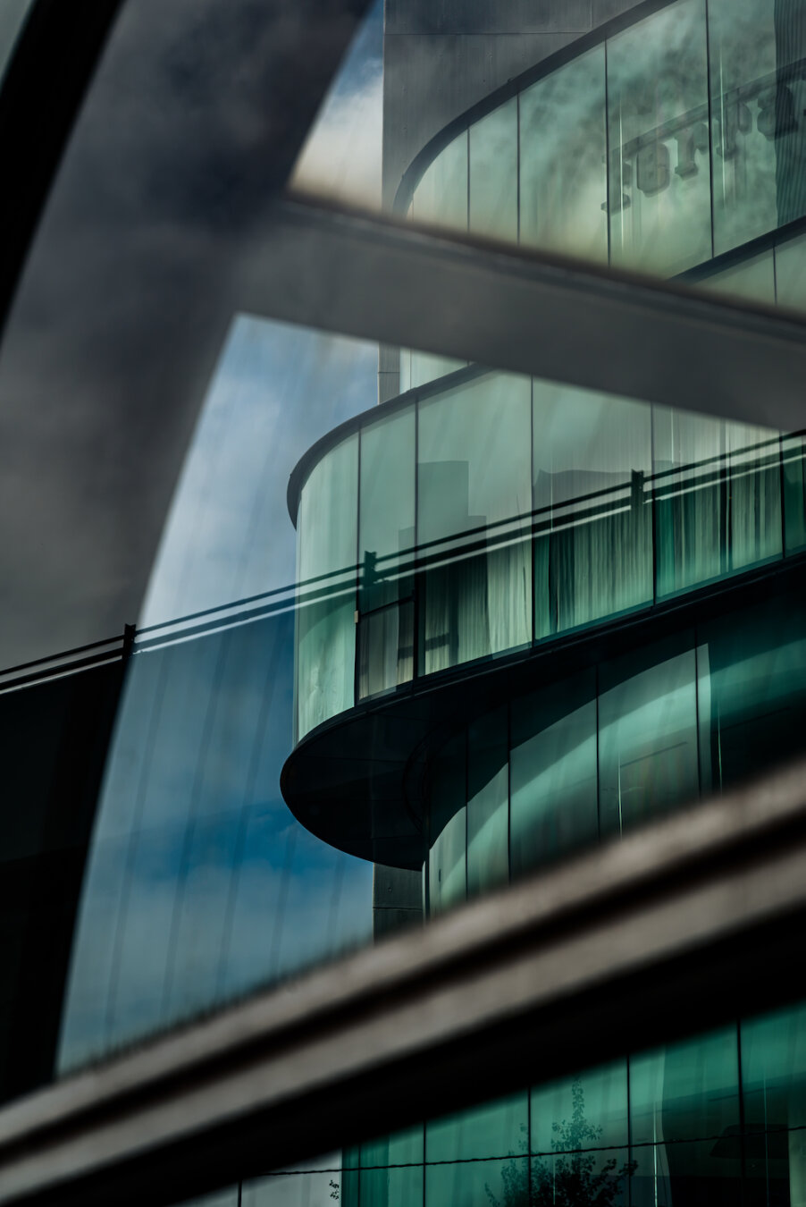 Viewed through the glass structure of an underground pedestrian entrance in Roppongi 5-chome, the curved glass facade of a Ferrari dealership creates a striking geometric reflection. Diagonal frames intersect the view, blending clouds and sky into the sleek emerald building surface, capturing a moody architectural contrast of light and shadow.