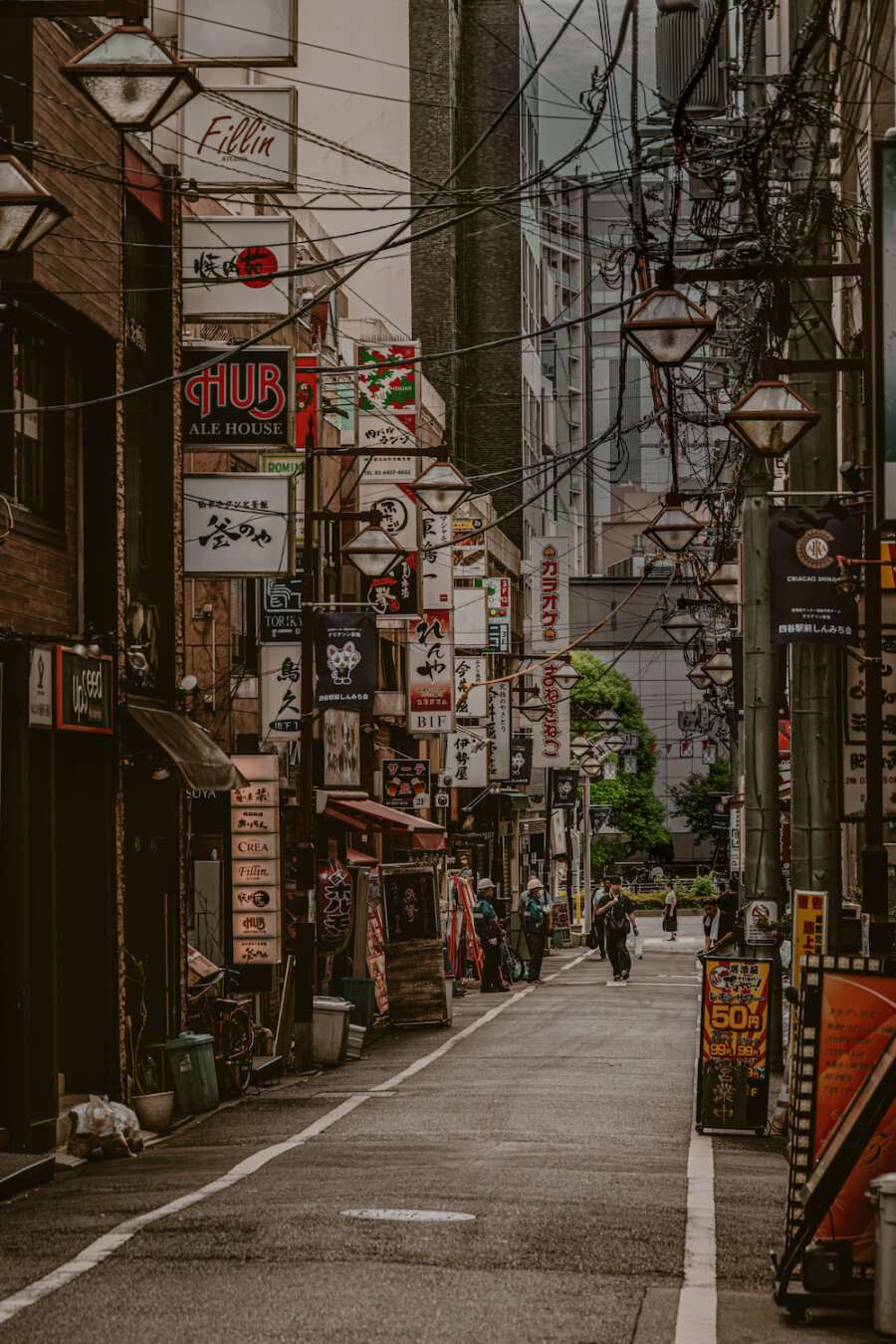 A quiet morning scene on Yotsuya Shinmichi-dori street in Tokyo. The narrow alleyway is lined with dense store signboards, lanterns and overhead cables, creating a nostalgic atmosphere. Only a few figures walk along the paved path, resting under the muted light before the busy day begins.