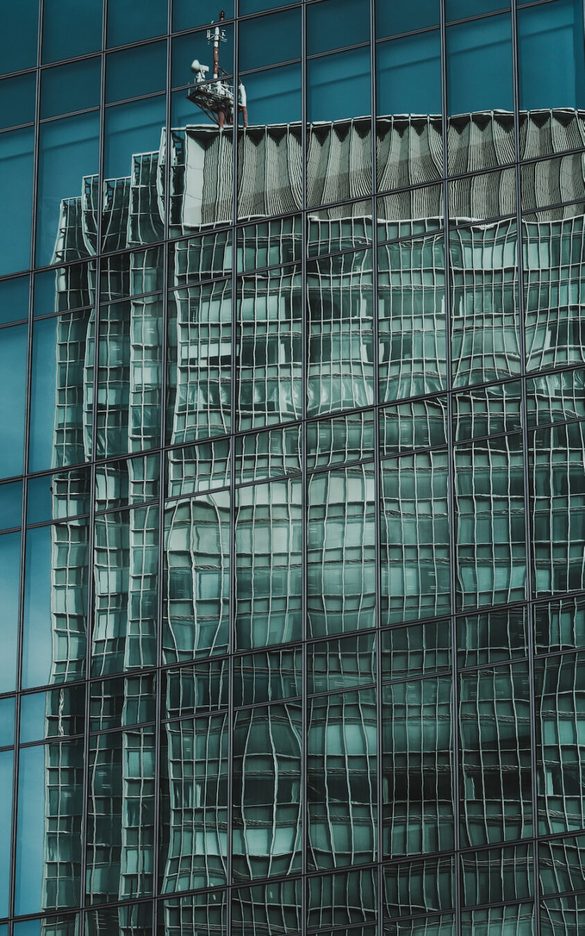 A towering glass facade reflects a neighbouring skyscraper and a lone antenna against a vibrant blue sky. This reflection is captivatingly distorted, transforming the precise grid of the urban landscape into fluid, wavy patterns. The shimmering lines form a surreal geometry within the smooth, blue panels, blending modern structure with dreamlike fluidity.