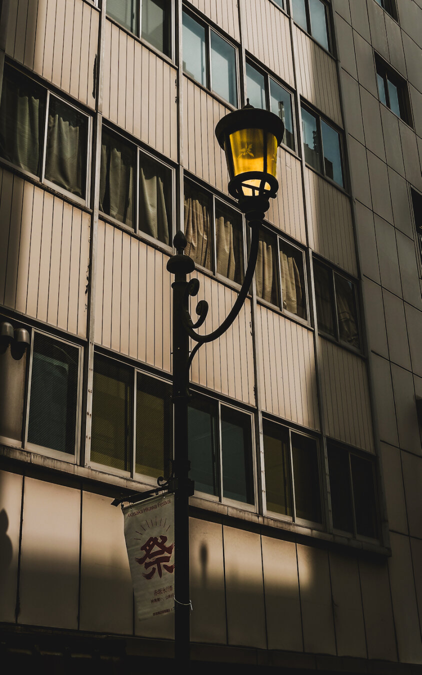 A nostalgic scene on Esplanade, where a historical building's facade is bathed in the warm, low light of the setting sun. The golden hour casts long shadows, illuminating a traditional, lit streetlamp in the foreground, creating a timeless atmosphere.