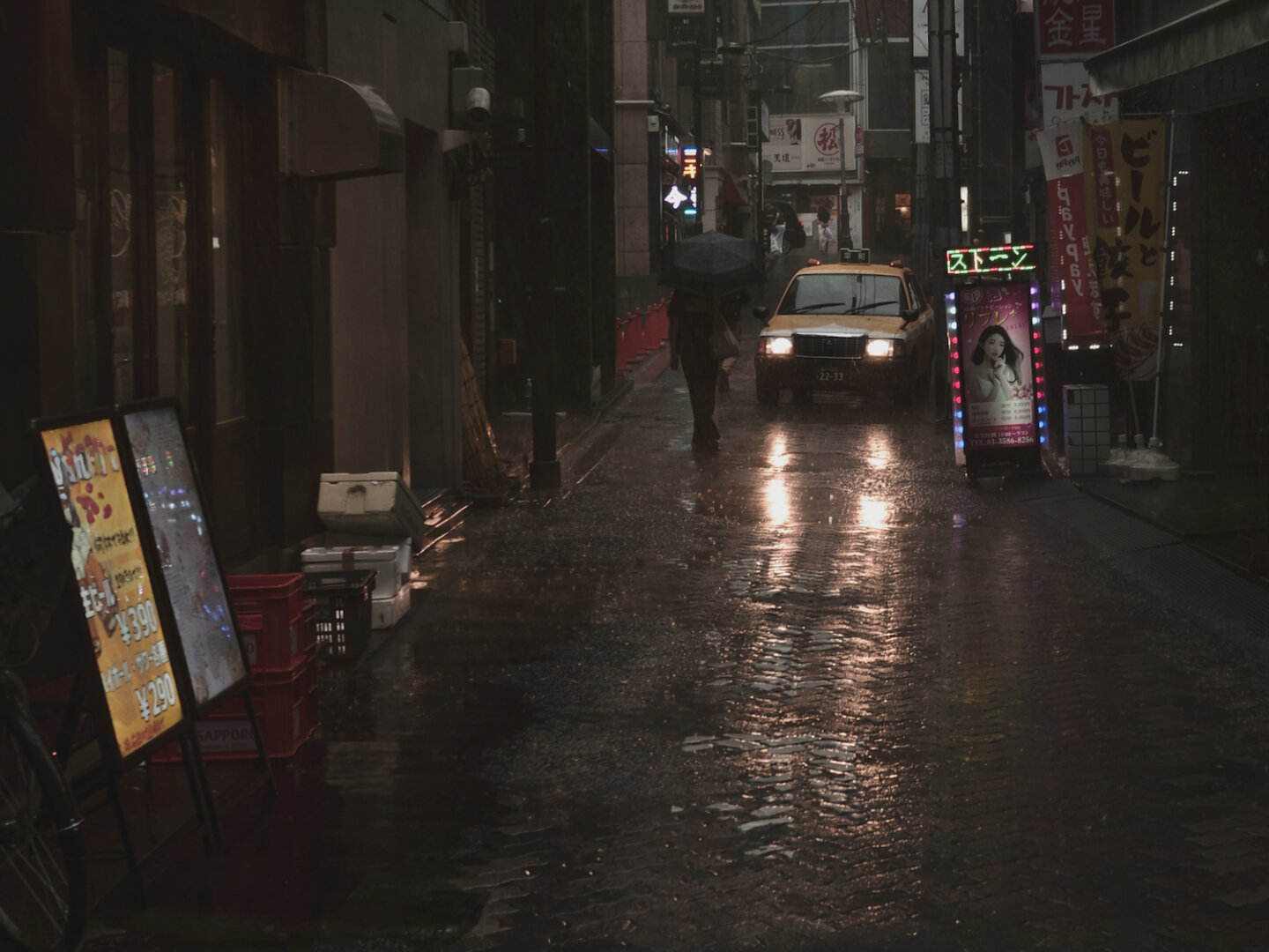 A yellow taxi slowly navigates a narrow, rain-slicked alleyway at night, its headlights casting long reflections on the wet asphalt. Nearby, a figure under a black umbrella walks past glowing neon shop signs, creating a dark, atmospheric urban scene filled with subtle light and deep shadows.