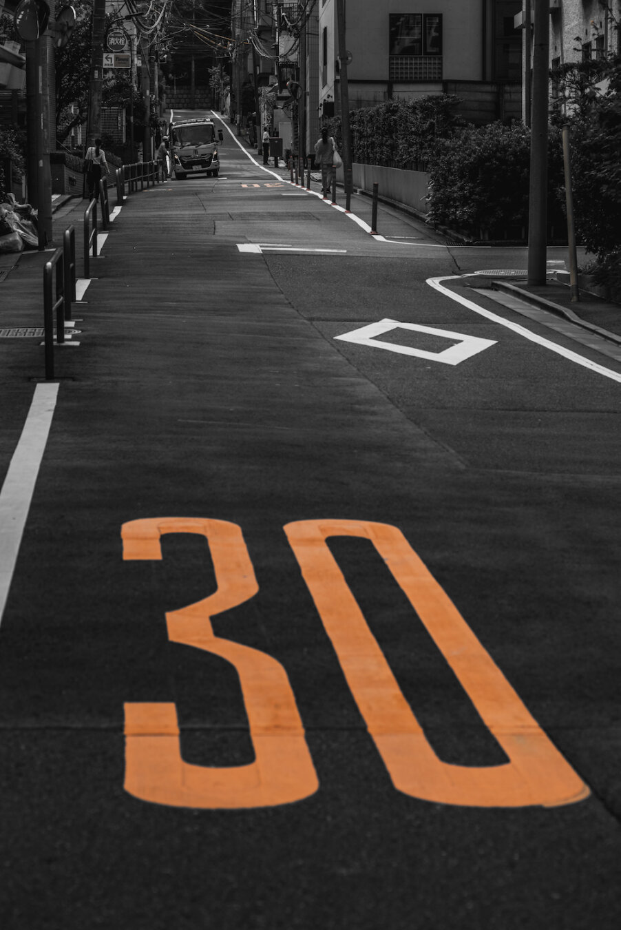 A monochrome urban street scene capturing Hikawazaka slope in Tokyo. In the foreground, a prominent orange speed limit mark '30' is painted on the asphalt. In the background, a refuse lorry ascends the steep incline, flanked by quiet residential buildings, creating a rhythmic harmony of daily urban routine.