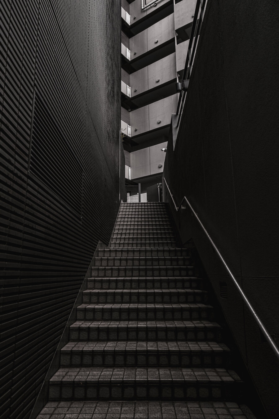 A vertical monochrome photograph looking up a steep, narrow, tiled staircase in an urban setting. Dark, textured, ribbed metal walls frame the path on the left and smooth dark concrete on the right with a metal handrail. In the distance, at the top of the steps, a multi-storey modern building with prominent balconies and concrete panels rises into the sky, its windows and railings creating patterns. The scene is dominated by shadow and strong leading lines, suggesting quietude and ascent.