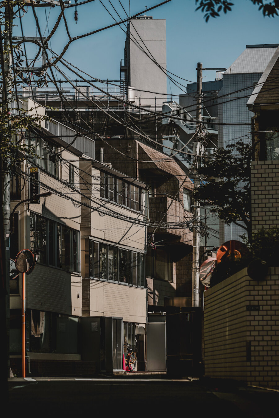A dense urban streetscape viewed from Inari Hill in Akasaka 7-chome, Tokyo. Intricate networks of overhead power lines criss-cross above geometric buildings, casting subtle shadows under the bright sky. The calm, shadowed slope leads through the quiet neighborhood, creating a striking contrast between modern infrastructure and peaceful architectural lines.