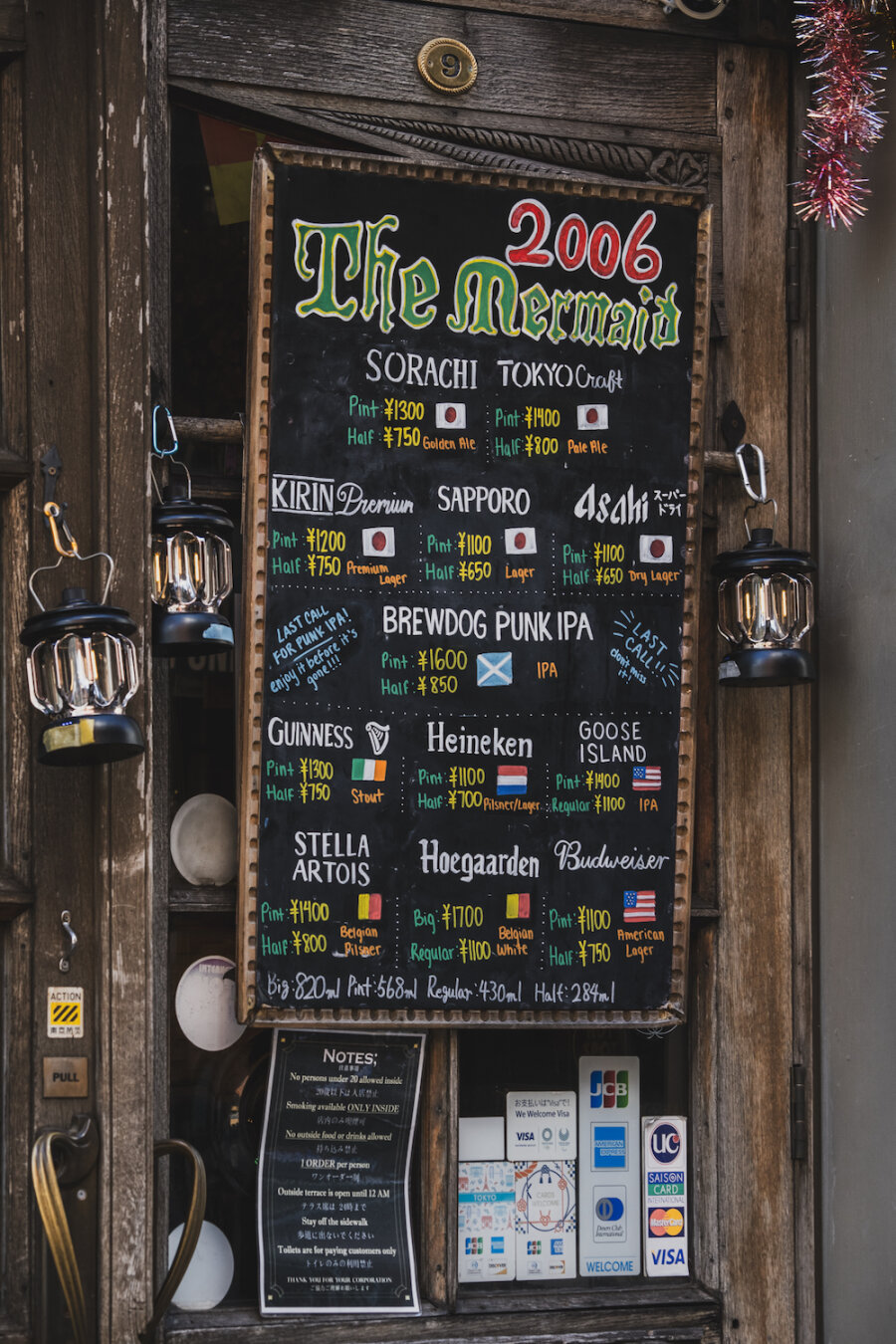 A hand-drawn chalkboard menu for 'The Mermaid' rests against an aged wooden door, framed by glowing vintage lanterns. Colourful lettering displays a varied beer selection, evoking a warm, nostalgic pub atmosphere. Below, small notes and payment signs anchor the rustic scene, inviting weary travellers into a cozy haven for the evening.