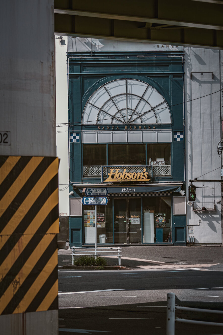 A calm street scene framed by a concrete flyover structure. Beyond the junction, the deep teal facade of the Hobson's store features a large semicircular arched window inscribed with SANTA BARBARA. Road signs for Roppongi-dori Ave. and Gaiennishi-dori Ave. stand quietly under the gentle afternoon sunlight.