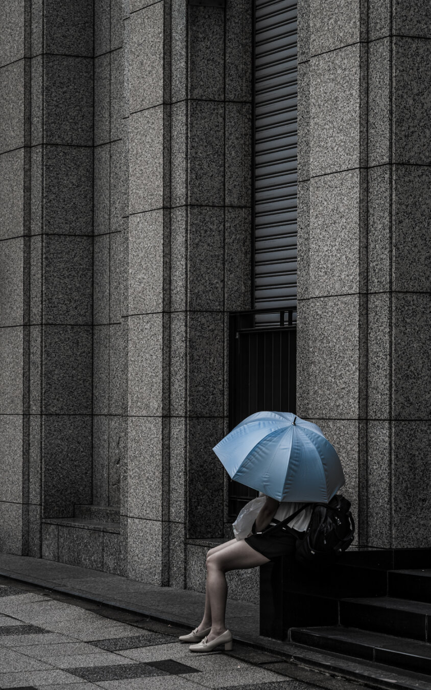 A minimalist portrait of a woman sheltered beneath a pale blue parasol. She sits in contemplation against a towering facade of grey geometric concrete, her light blue sanctuary providing contrast to the sterile, monochromatic architecture. She is a momentary, quiet accent in the immense urban sprawl.