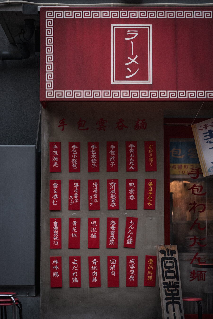 A traditional ramen shop facade displays a bright red awning above a concrete wall adorned with vibrant red menu cards in calligraphic script. One card space is noticeably missing from the neat grid, leaving a quiet vacant spot that creates a subtle, nostalgic narrative within the street scene.