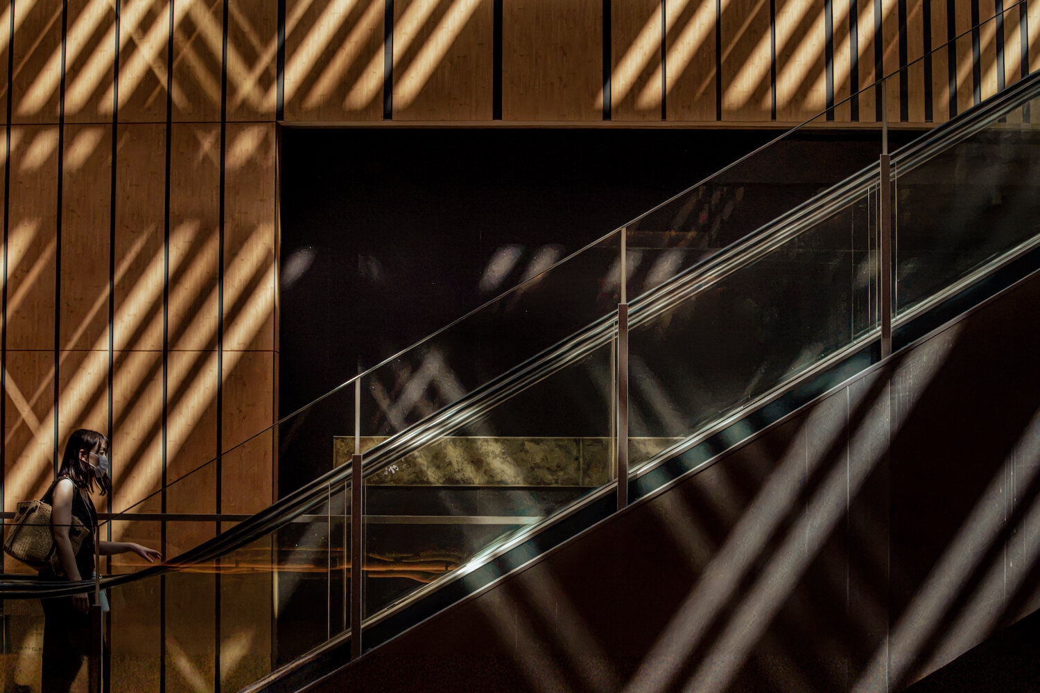 Dramatic diagonal stripes of sunlight and shadow stretch across a wooden wall and an escalator. A solitary woman in a face mask moves along the handrail, bathed in the rhythmic geometric light that transforms a modern architectural space into a poetic interplay of brilliance and shade.