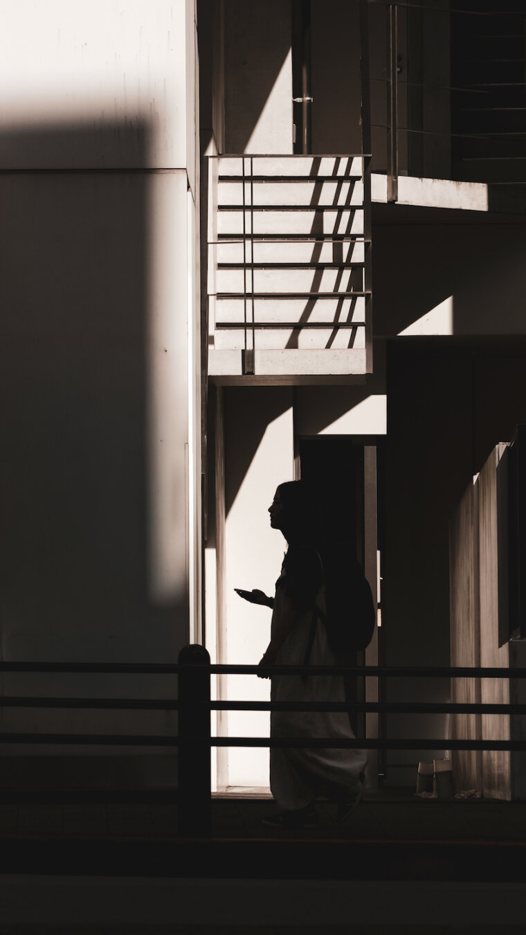A stark, high-contrast urban photograph showing a person in dark silhouette walking past modern architectural structures. Bold sunlight casts strong geometric shadows across light-coloured concrete walls and balconies. The figure, holding a mobile device, moves gracefully through the sharp interplay of light, shadow and strong structural lines.