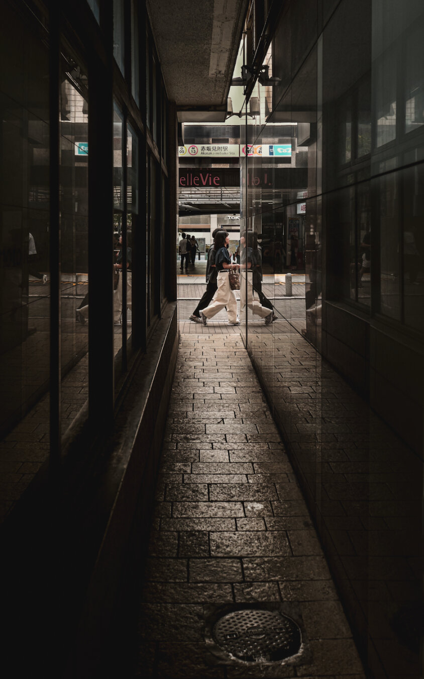 A narrow, dark alley framed by glassy reflections opens onto a sunlit street near Akasaka-mitsuke Station in Tokyo. Pedestrians walk past the threshold, captured mid-step where shadow meets bright urban life, creating a quiet contrast between the secluded passage and the bustling city beyond.