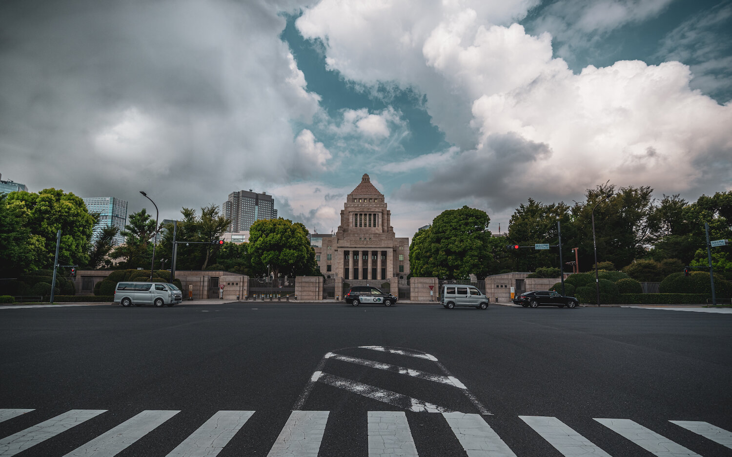 Beyond a vast tarmac road with zebra crossings, the National Diet Building stands resolutely. Dark, dynamic storm clouds gather aggressively in the sky above the granite structure and dense green trees, while a single grey minivan is parked centrally. A dramatic contrast plays out between the enduring architecture and the rapidly shifting elements.