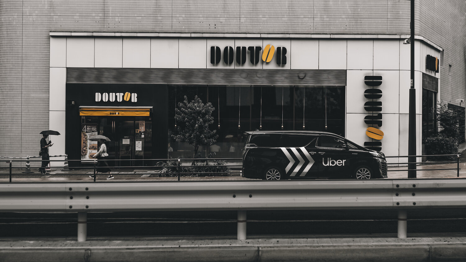 A black vehicle with an Uber logo passes a Doutor Coffee shop. Pedestrians walk by under umbrellas on a rainy day. Yellow coffee bean logos stand out amidst monochrome urban tones, creating a rhythmic dialogue of modern city life.