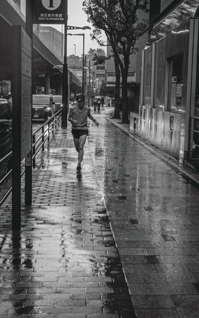 A monochrome street photograph capturing a solitary male runner in motion along a rain-slicked pavement in Minato City, Tokyo. His determined figure is mirrored in the wet surface, moving forward under the gaze of a Minato City Information Map sign and against a backdrop of traffic and urban architecture blurring in the heavy downpour.
