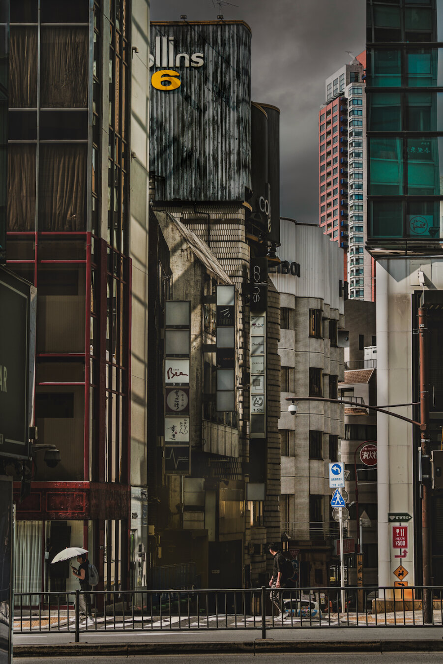 A dramatic street view in Roppongi, Tokyo, where the old Collins 6 building stands. Its distressed texture is sharply illuminated next to a modern skyscraper. Deep shadows veil a solitary figure with an umbrella and another walking. Light cuts through the urban canyon, creating a stark, poetic contrast under a grey sky.