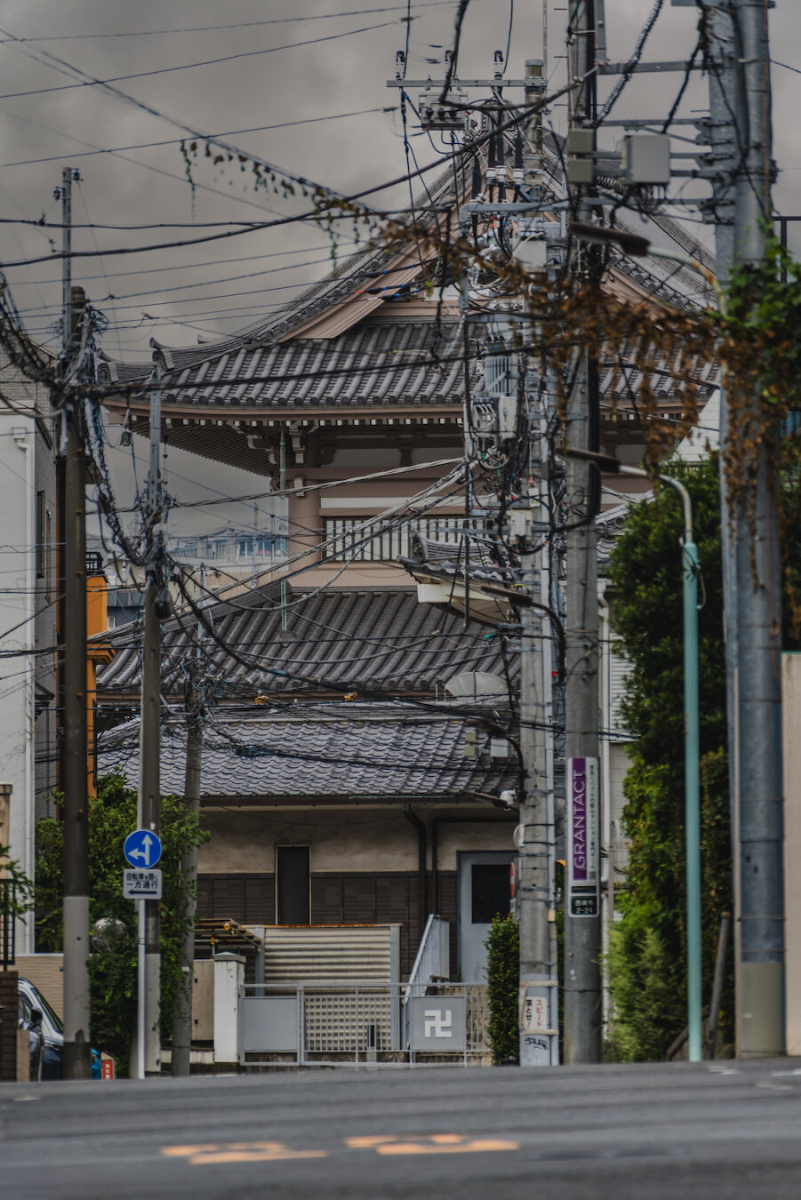 Beyond a web of overhead power lines strung along Nishiazabu's urban landscape, an ancient Buddhist temple pagoda is glimpsed. The traditional multi-tiered roof rises above the wires and nearby greenery, creating a striking contrast between old and new Tokyo under a dramatic cloudy sky.