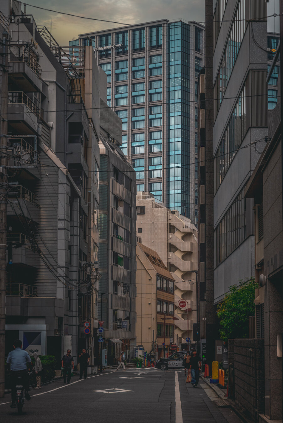 Looking down a shadowed narrow street framed by dense urban buildings in Kojimachi, Chiyoda. At the end of the dim passage, a massive glass high-rise towers dramatically under the overcast sky, catching the soft ambient light and standing as a glowing beacon above the quiet city canyon below.