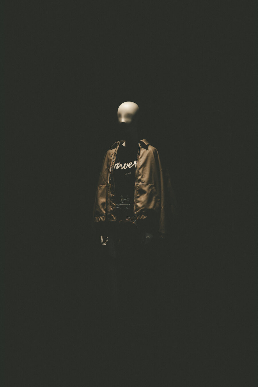In the quiet morning light of Omotesando, Minami-Aoyama 6-chome, a solitary mannequin stands within a darkened shop display. Wearing a stylish jacket over a graphic top, its smooth, featureless form emerges softly from deep shadows, capturing a peaceful moment of urban stillness before the city awakens.