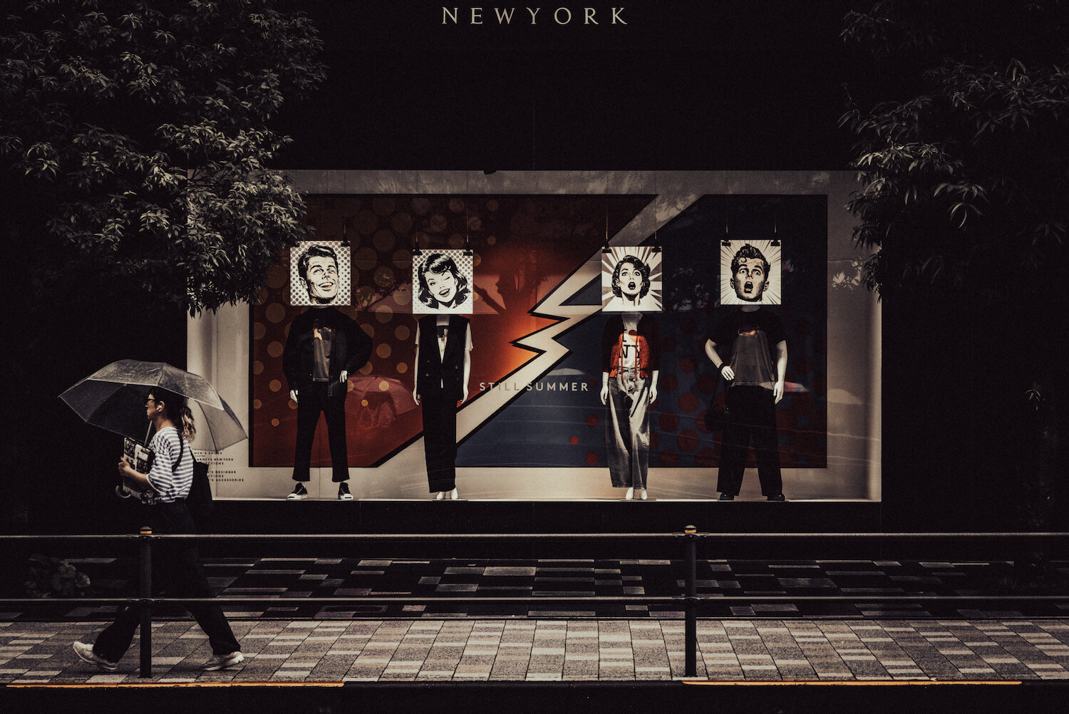 A person holding a clear umbrella walks past the stylish window display of Barneys New York Roppongi on a rainy day. Behind the glass, mannequins with pop-art comic face illustrations stand before a bold lightning bolt graphic bearing the phrase STILL SUMMER, creating a striking contrast between daily urban life and artistic fashion.