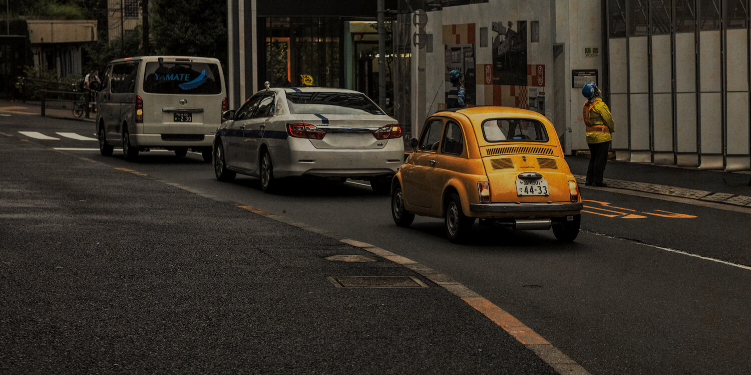 From behind, a vintage, vibrant yellow classic car cruises down a multi-lane Tokyo street, its small frame contrasting with modern vehicles. A silver van and a white saloon car are in front. Behind the yellow car, a person in a hi-vis jacket stands by the kerb, under a muted urban sky.
