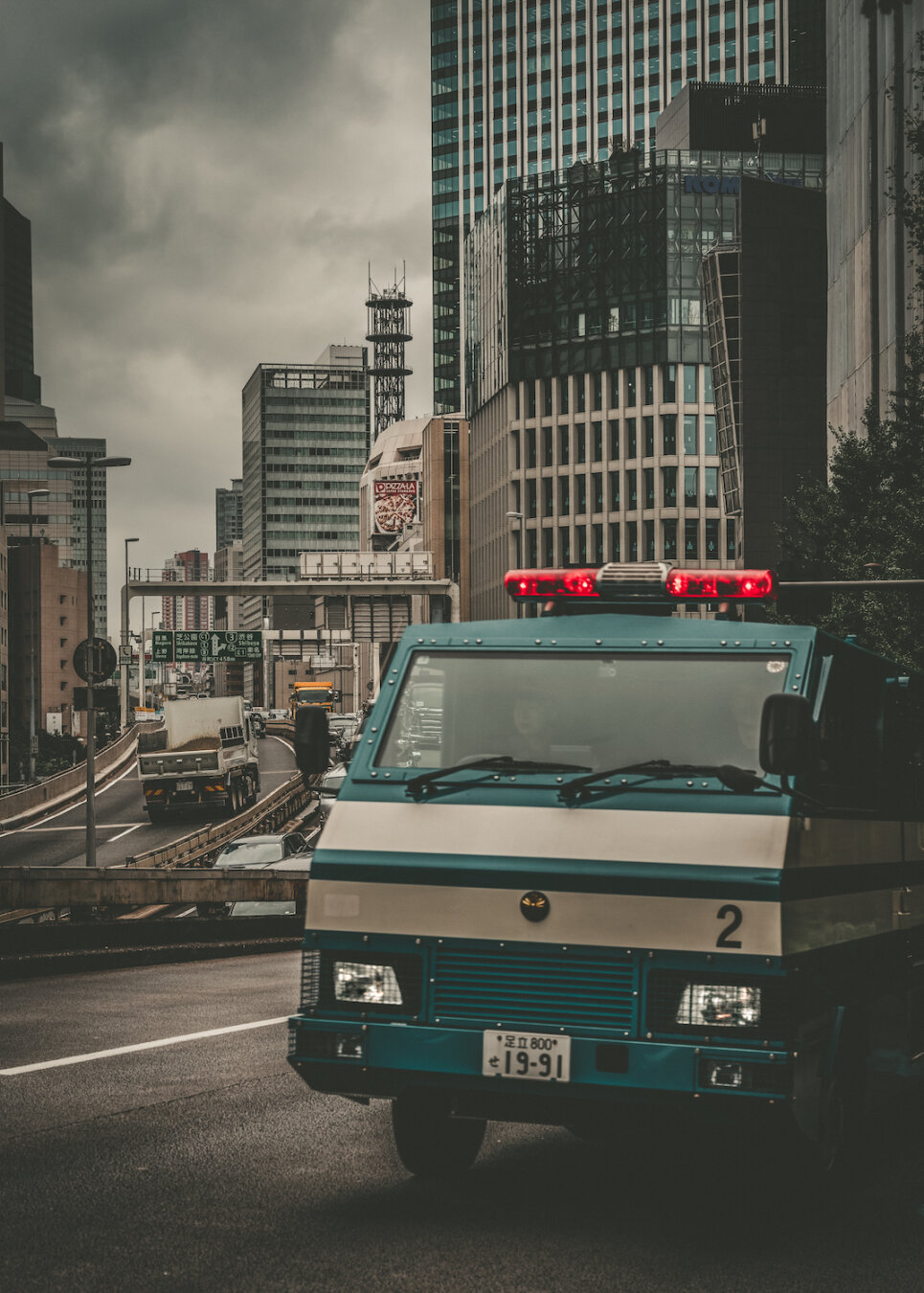A dark green police security vehicle with flashing red lights drives along a street in Tameike-sanno, Tokyo. Modern high-rise glass buildings and an elevated expressway loom beneath a heavy grey sky, creating a quiet yet tense atmosphere in the urban landscape.