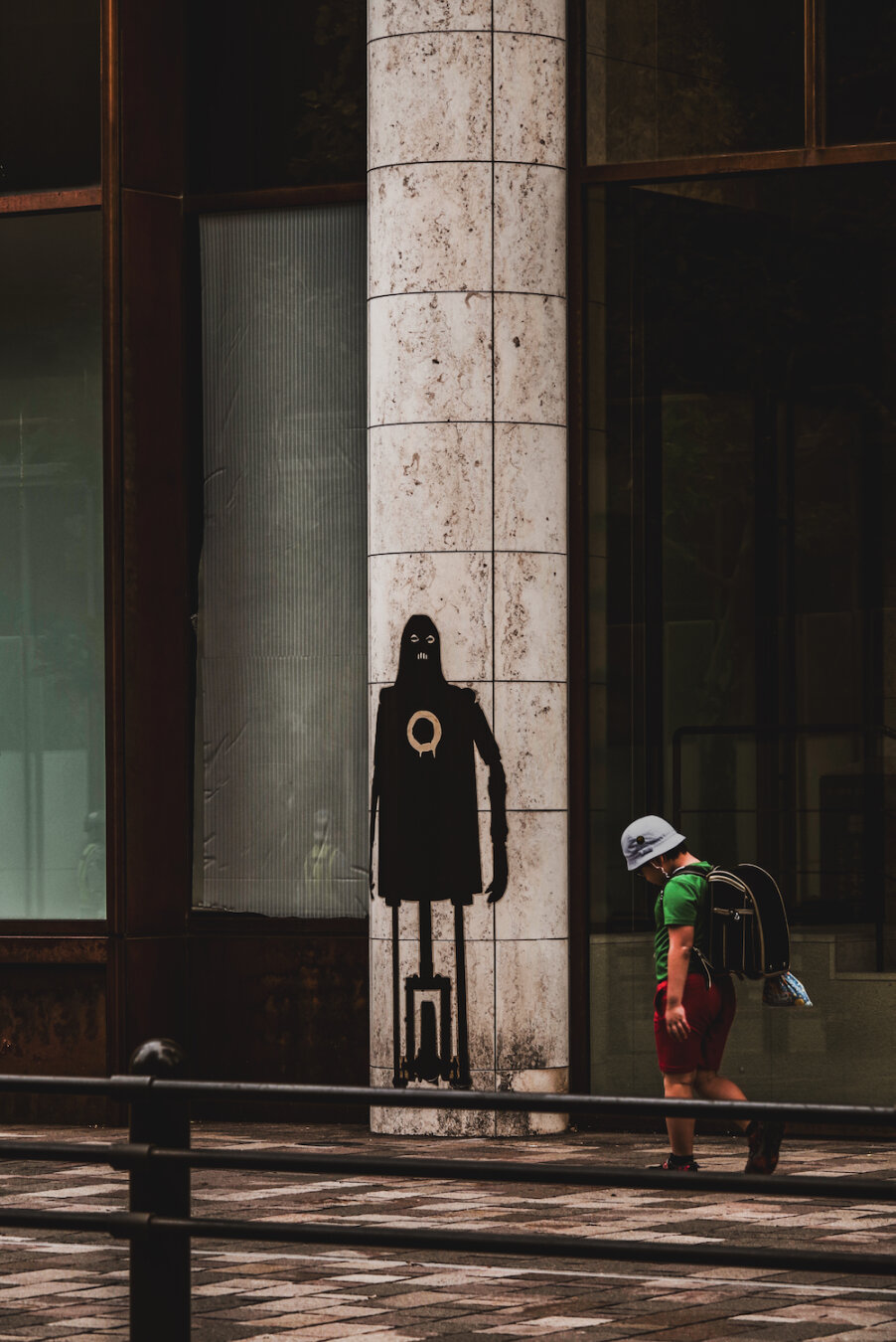 A young student wearing a backpack walks past a stone pillar featuring a dark, mysterious mural figure. Standing as a silent guardian along the pavement, the painted character on the column softly watches over the child's daily commute, blending subtle street art with a quiet moment of everyday urban life.