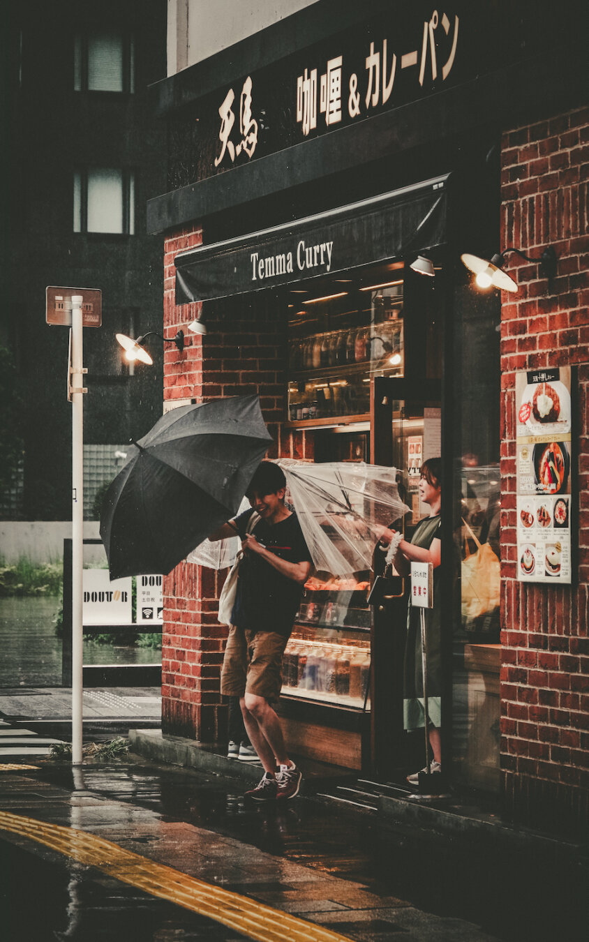 At the entrance of Temma Curry, a traditional brick-walled restaurant, people open umbrellas and step onto a rain-slicked Tokyo street. The warm glow from the shop contrasting with subtle shadows and reflective textures captures a nostalgic urban moment under soft daylight, free of any file names.