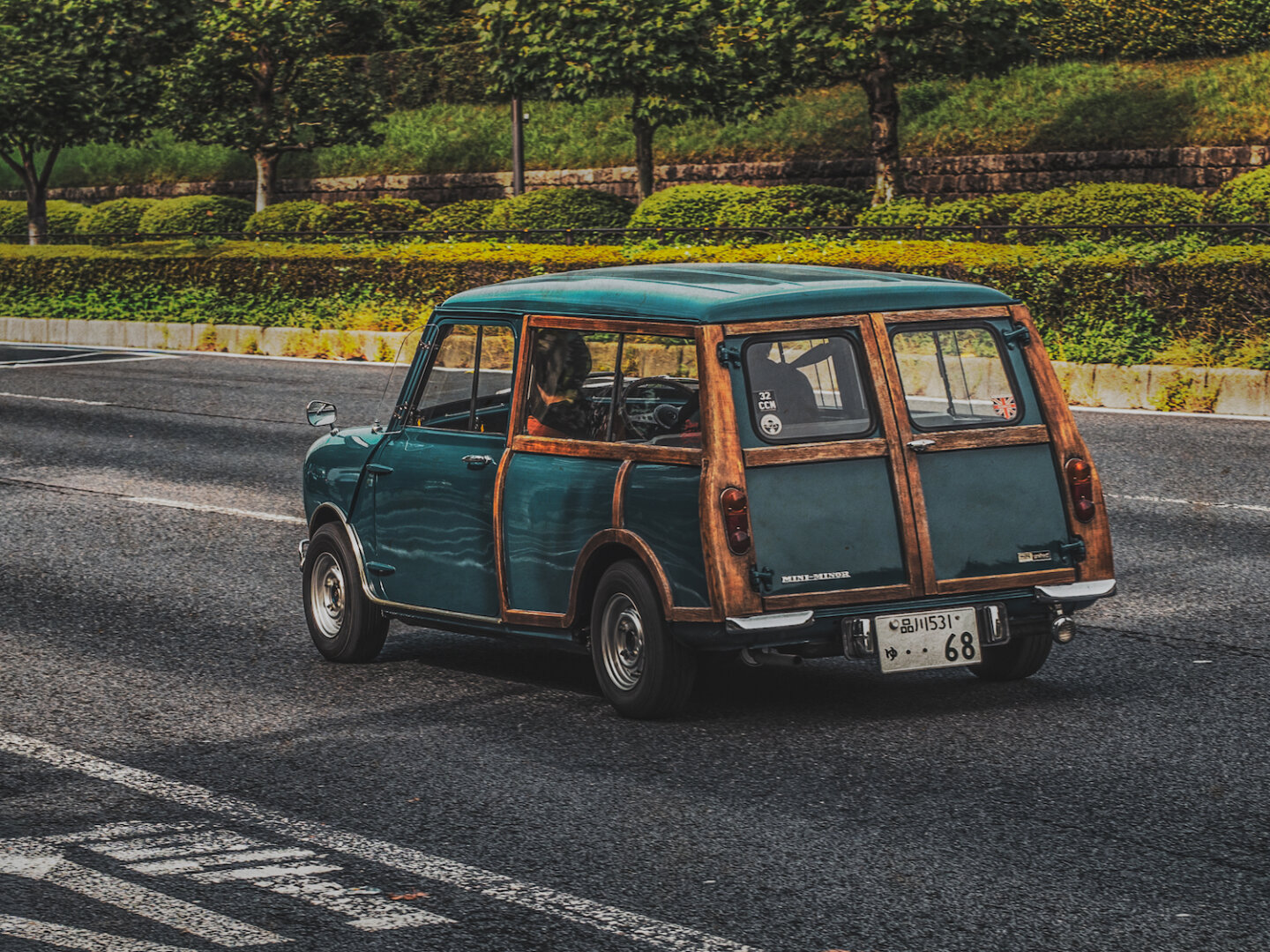 A vintage green Rover Mini with classic wooden framing cruises gracefully along an asphalt road. Warm sunlight glints off its metallic body and rich timber trim, creating a nostalgic scene set against neatly manicured roadside greenery.