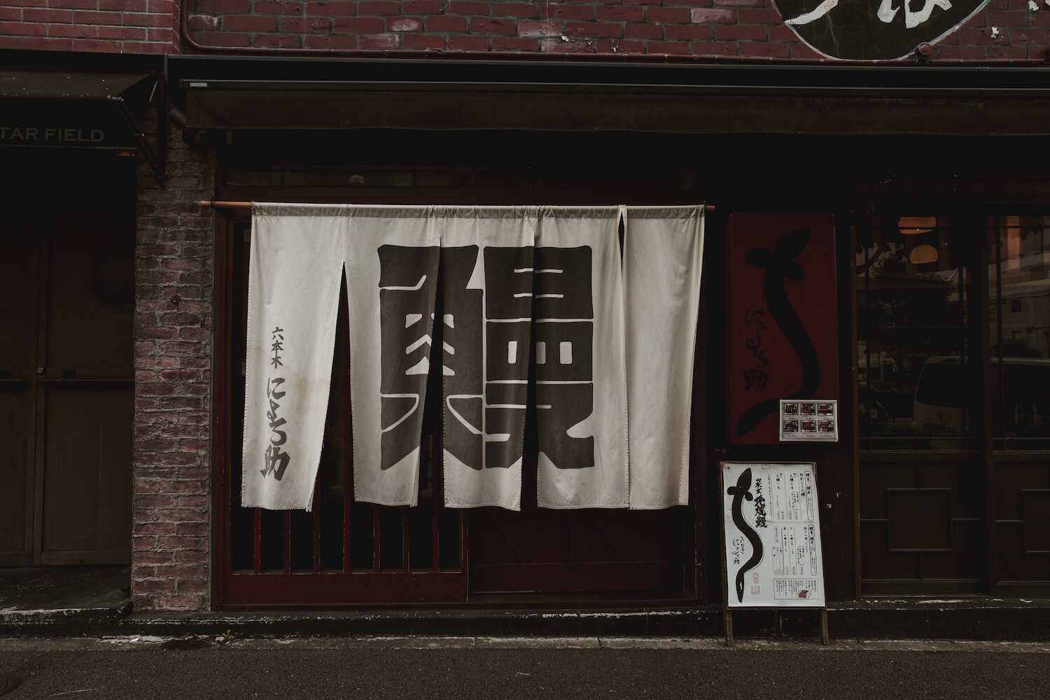 A clean white fabric noren curtain hangs outside a traditional unagi (eel) restaurant, its surface bearing the calligraphic Japanese character '鰻' (unagi) meaning eel. Set against weathered dark wood panels and red accents, this serene street-front scene in Tokyo invites passersby into the deep, fragrant tradition of grilled freshwater eel.