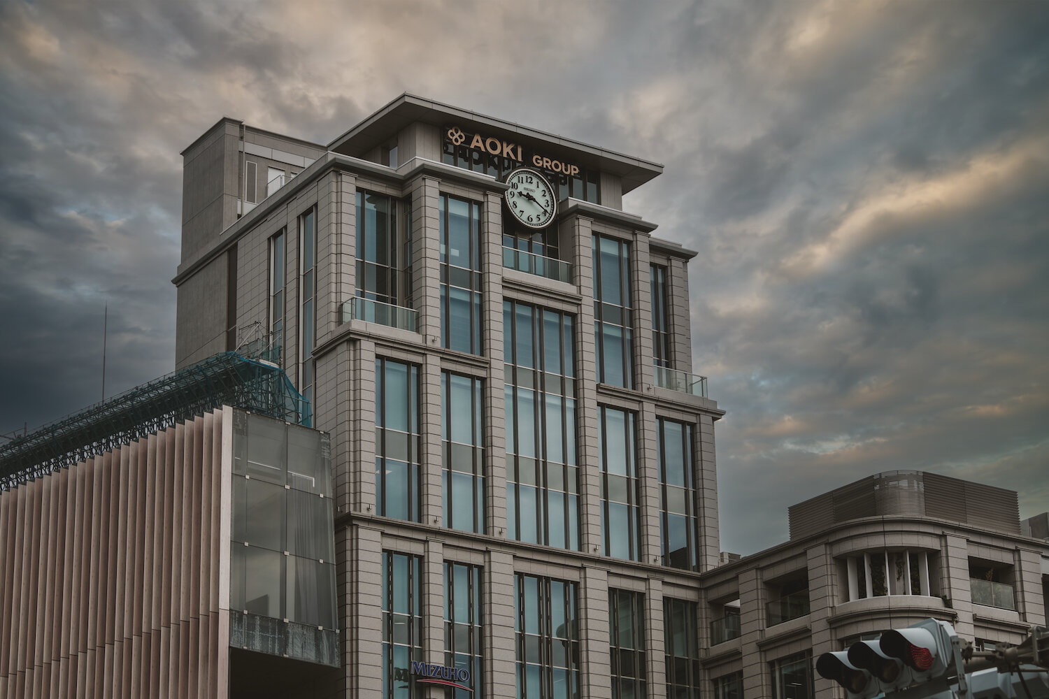 A modern building with stone cladding stands in Omotesando, Tokyo, under a dramatic cloudy sky. At the top, a large clock reads twenty past nine. Below the AOKI GROUP sign, the structure rises elegantly, blending classic architectural dignity with dramatic urban atmosphere.