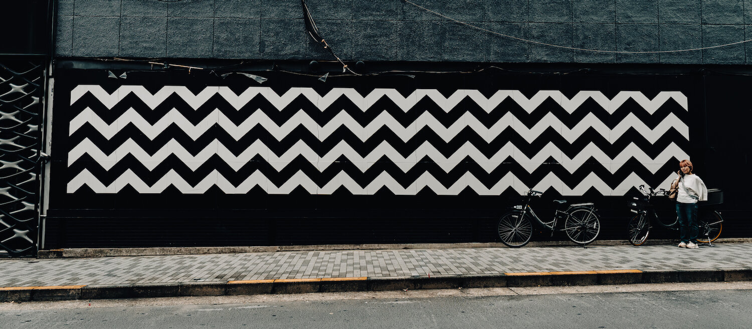 A striking black and white mural with a bold zigzag pattern spans a dark wall along a quiet street in Higashi-Azabu. Two bicycles are parked beside the rhythmic graphic artwork, where a person stands quietly, creating a captivating visual balance between modern geometric street art and the subtle calm of daily urban life.