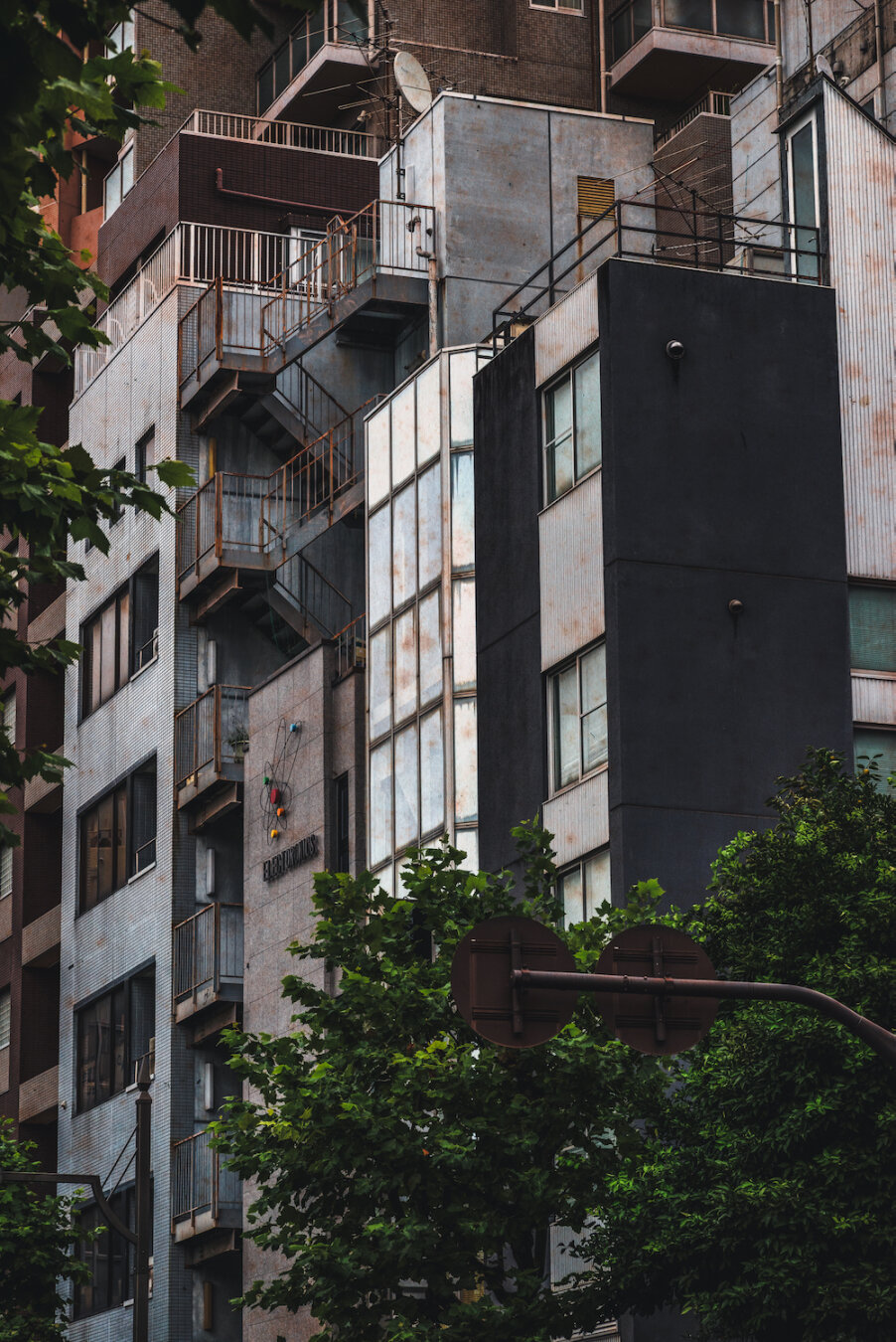 Slender, weathered buildings stand side by side in a dense urban landscape, their rusty emergency stairs, aged metallic panels, and glass facades rising quietly above lush green street trees, weaving a silent story of city life.